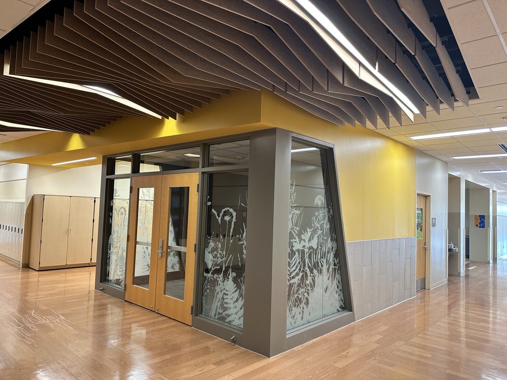 Finished stepped, faceted Soundcore baffle ceiling in English Walnut above a yellow-accented breakout room entrance at Westwood Elementary School, with a matching nature-themed frosted glass graphic